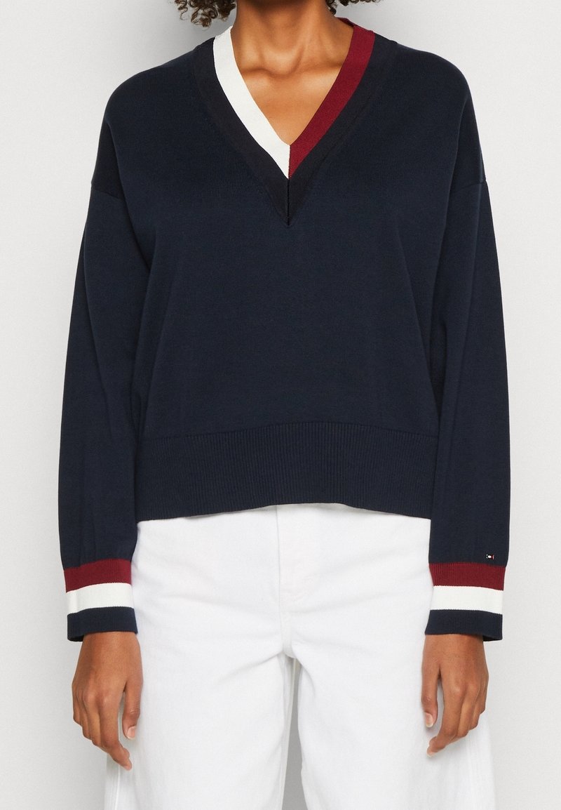 Pull bleu marine avec encolure en V blanche et bordeaux et poignets rayés assortis porté avec un pantalon blanc.