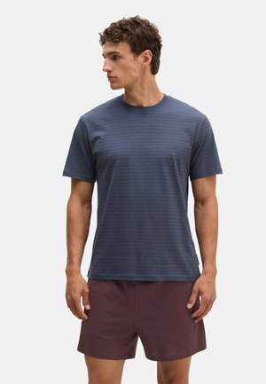 Mann mit kurzen lockigen Haaren, der ein blau gestreiftes Kurzarmhemd und bordeauxrote Shorts trägt, blickt nach links vor einem einfarbigen Hintergrund.