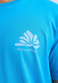 Modré tričko s krátkým rukávem vyrobené z bavlny, s bílým logem se stylizovanými okvětními lístky a textem "Petrol®Ind" na přední straně.