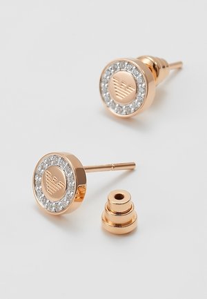 Pendientes de botón en oro rosa con borde de cristal y logo de águila grabado, mostrados con el soporte de un pendiente sobre un fondo blanco.