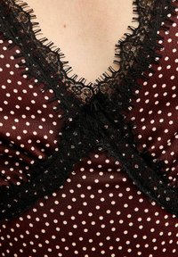 Tejido burdeos con lunares blancos, presenta un borde de encaje negro alrededor del escote y un detalle de volantes en el borde. Textura suave, diseño ajustado.