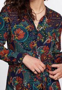 Kvinna som knyter midjebältet på en långärmad skjortklänning med flerfärgade blommönster och geometriska mönster, bärande ett guldhalsband och ringar.