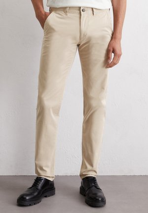 Beige cotton trousers with a slim fit, five-pocket design, and visible stitching. Paired with black leather shoes, standing on a gray surface.