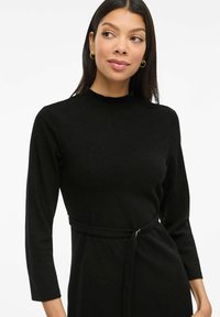 Black long-sleeved dress with a high neckline, belted waist, and soft texture. Simple design with minimal accents and a fitted silhouette.
