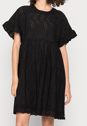 Frau trägt ein locker sitzendes schwarzes Kleid aus Lochstickerei-Stoff mit gerafften kurzen Ärmeln und einer betonten Taille, dargestellt von den Schultern bis zu den Knien.