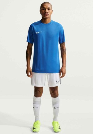 Mężczyzna stojący, ubrany w niebieską sportową koszulkę z krótkim rękawem, białe szorty i skarpetki oraz neonowożółte korki z czarnymi logo Nike.