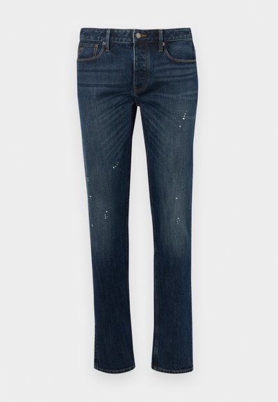 Mørkeblå smale jeans med lett falming og små hvite malingssprut, med fem lommer og knappelukking.