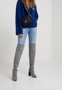 Graue, karierte, oberschenkelhohe Stiefel mit einem lockeren Design und Blockabsätzen, getragen zu hellblauen Jeans und einem blauen Strickpullover sowie einer schwarzen Crossbody-Tasche.