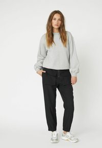Felpa grigia in cotone con polsini a coste, abbinata a pantaloni neri tapered con tasche frontali. Il modello indossa sneakers bianche e argentate.