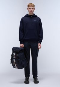 Marine hoodie met een voorzak, gecombineerd met donkere broeken en zwarte schoenen. Een grote marine rugzak met grafische accenten wordt in de linkerhand vastgehouden.