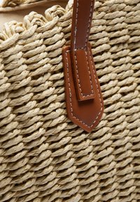 Sac en paille tressée avec une anse en cuir marron ornée de coutures blanches et une surface texturée. Le design est circulaire avec une palette de couleurs naturelles.