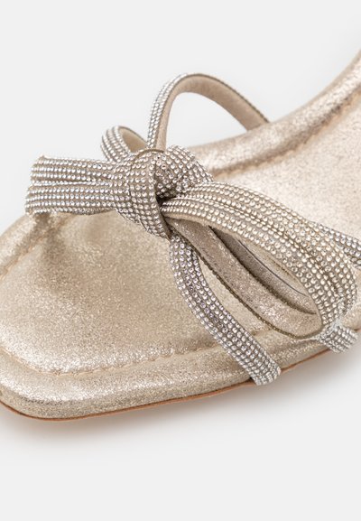 Sandale dorée avec une finition métallique, présentant une sangle nouée ornée de détails en argent texturé et un design à bout carré.