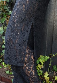 Fekete csipke anyag virágmintákkal, magas hasítékkal. Az anyag finom kidolgozású, finom mintázatot mutat sötét háttéren.