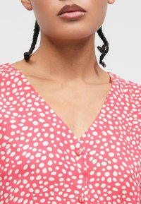 Ροζ φόρεμα με λευκές κουκίδες, V-λαιμόκοψη και κοντά μανίκια, με ροζ κουμπιά μπροστά και λεία υφή υφάσματος.