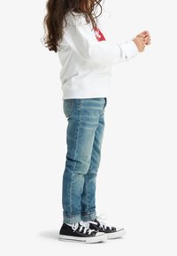 Vit sweatshirt med röd logotyp, blå jeans och svarta sneakers med vita sulor. Jeansen är uppvikta vid fållarna, avslappnad passform. Håret är vågigt.