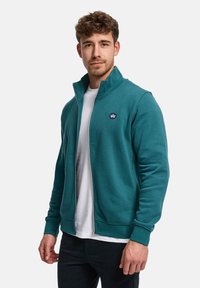 Petroleumsfarget hettegenser med glidelås laget av mykt materiale, med høy krage og ribbestrikkede mansjetter, lagd over en hvit t-skjorte. Liten logo på brystet.