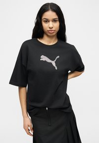 Νεαρή γυναίκα με μακριά σκούρα μαλλιά φοράει μια φαρδιά μαύρη μπλούζα Puma με λογότυπο από στρας και μια μαύρη φούστα, μπροστά σε απλό λευκό φόντο.