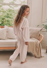 Set di pigiama in tessuto rosa tenue, composto da un top a maniche lunghe ampio con arricciature e pantaloni con polsini elasticizzati. Stanza con texture accoglienti e piante.