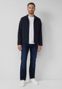Marineblaues Zip-Jackett über einem weißen T-Shirt, kombiniert mit dunkelblauen Jeans und weißen Sneakern; glatter Stoff, schlanker Schnitt, schlichtes Design.