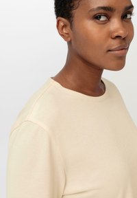 Helles beige T-Shirt mit kurzen Ärmeln und Rundhalsausschnitt. Weicher, glatter Stoff mit dezenten Nahtdetails an den Schultern.