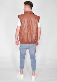 Brun læder ærmeløs jakke med krave, tekstureret overflade og elastisk kant, parret med lys blå jeans og hvide sko.