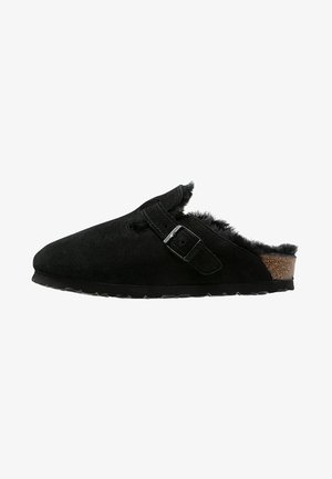 Zwarte leren klompen met een enkele verstelbare gesp, zachte fuzzy voering en een gestructureerde rubberen zool voor grip en comfort.