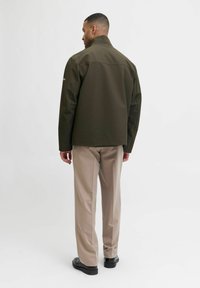 Temno olivno nepremočljivo jacket z usnjeno ovratnikom in minimalističnim dizajnom, v kombinaciji s svetlo bež hlačami in črnimi čevlji.