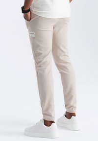 Pantalones de chándal beige en un ajuste entallado, con una tela texturada, bolsillos laterales y una etiqueta en el muslo izquierdo, combinados con zapatillas blancas.