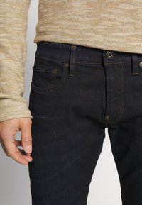 Muž v tmavě modrých džínách a béžovém svetru s dlouhým rukávem, ukazující pas, kapsy a levou ruku uvolněně podél těla.