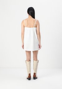 White sleeveless dress with adjustable straps and gathered back, paired with knee-high white and brown cowboy boots featuring decorative stitching.