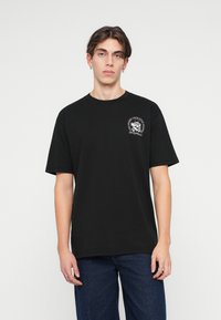 T-shirt de algodão preto com decote redondo e mangas curtas. Apresenta um gráfico em branco na parte da frente, no peito, com detalhes intrincados.