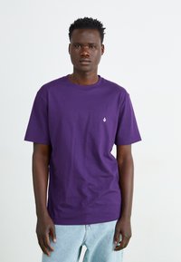 T-shirt en coton violet avec un col rond, des manches courtes et un petit logo blanc sur la poitrine gauche. Porté avec un jean bleu clair.