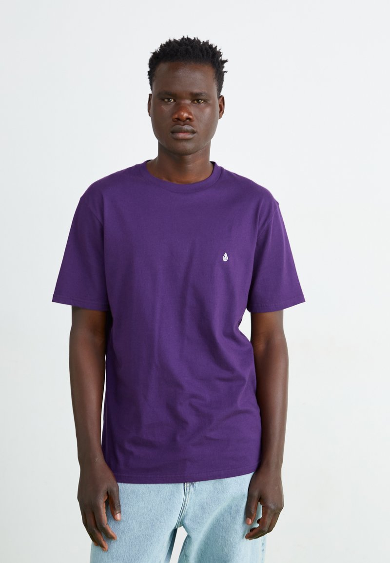 T-shirt en coton violet avec un col rond, des manches courtes et un petit logo blanc sur la poitrine gauche. Porté avec un jean bleu clair.
