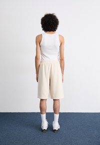 Calças de bermuda em cor creme com um cós elástico, combinadas com uma t-shirt branca. O fundo é branco liso e o chão é azul.