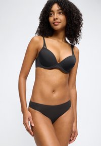 Γυναίκα με σγουρά μαλλιά που φοράει μαύρο σουτιέν με διπλούς ιμάντες στους ώμους και ταιριαστή μαύρη εσώρουχα, στέκεται μπροστά σε απλό λευκό φόντο.