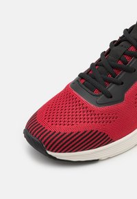 Close-up of red and black knit running shoe with black laces and white sole, highlighting textured fabric and toe area.