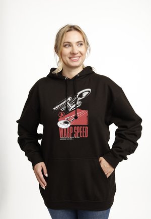 Smilende kvinde med blondt hår iført en sort hoodie med et billede af et rumskib og rød tekst, der lyder "Warp Speed". Hænder i lommerne, hvid baggrund.