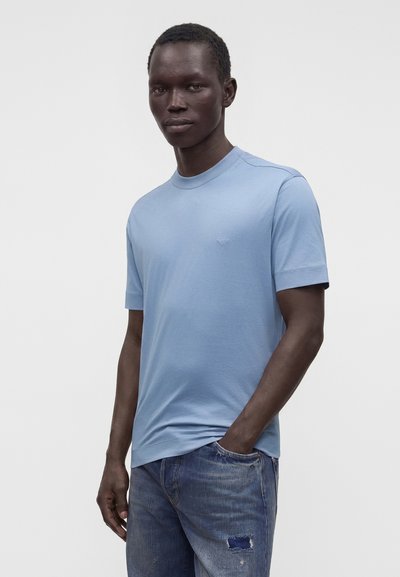 Άνδρας που φοράει στενό, ανοιχτό μπλε t-shirt με κοντά μανίκια και μπλε τζιν με ένα μικρό μπαλώμα, στέκεται με το ένα χέρι στην τσέπη.