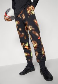 Black sweatpants with a vibrant abstract pattern in orange and green; tapered fit and ribbed cuffs. Holding a black and white soccer ball.
