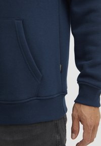 Marinblå sweatshirt med en struktureradtyg, ribbade mudd och nederkant, känguruficka och en liten märkesetikett på sidan.