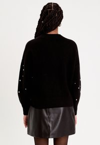 Pull noir à col rond, avec des accents de perles le long des manches. Associé à une mini-jupe en cuir noir.