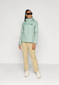 Šviesiai žalia, neperšlampama striukė su pilnu užtrauktuku, reguliuojama kapišonu ir logotipo lipduku, derinama su smėlio spalvos kelnėmis ir šviesiais bateliais. Lygi tekstūra.