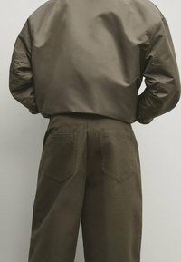 Bruine nylon bomberjack in combinatie met bruine katoenbroek, met twee achterzakken en een relaxte pasvorm, gezien vanuit een achteraanzicht.