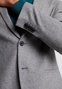 Närbild av en person som bär en grå ullblazer över en teal-färgad tröja, med ena armen korsad över bröstet.