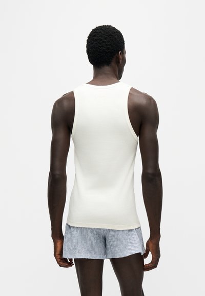 Homme à la peau foncée portant un débardeur blanc sans manches et un short rayé bleu clair, debout de dos sur un fond uni.