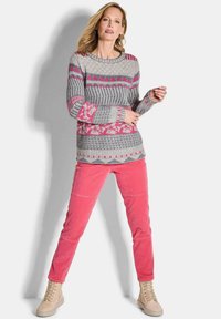 Eine Frau steht in einem gemusterten grauen und rosa Pullover, leuchtend rosa Hosen und beigen Schnürstiefeln vor einem schlichten hellen Hintergrund.