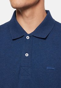 Temno modra polo majica z teksturirano tkanino, tremi gumbi in majhnim modrim poudarkom nad levim robom.