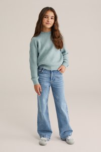 Šviesiai mėlynas raštuotas megztinis su ilgomis rankovėmis, derinamas su plačiomis mėlynomis džinsomis su subtiliais papuošimais. Dėvimos pilkos sportbačiai.