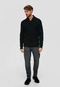 Zwarte gebreide trui met een sjawlboord, gecombineerd met donkere spijkerbroek en zwarte schoenen. De trui heeft een gestructureerde stof en lange mouwen.