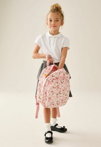 Jeune fille en polo blanc et jupe grise tenant un sac à dos rose fleuri, portant des chaussettes blanches et des chaussures noires, debout devant un fond uni.